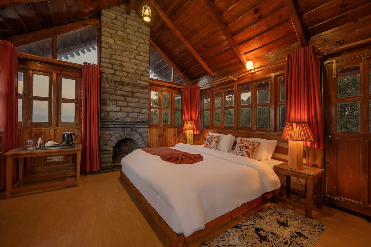 A cozy room with a large bed, wooden walls, a stone fireplace, and large windows with curtains.