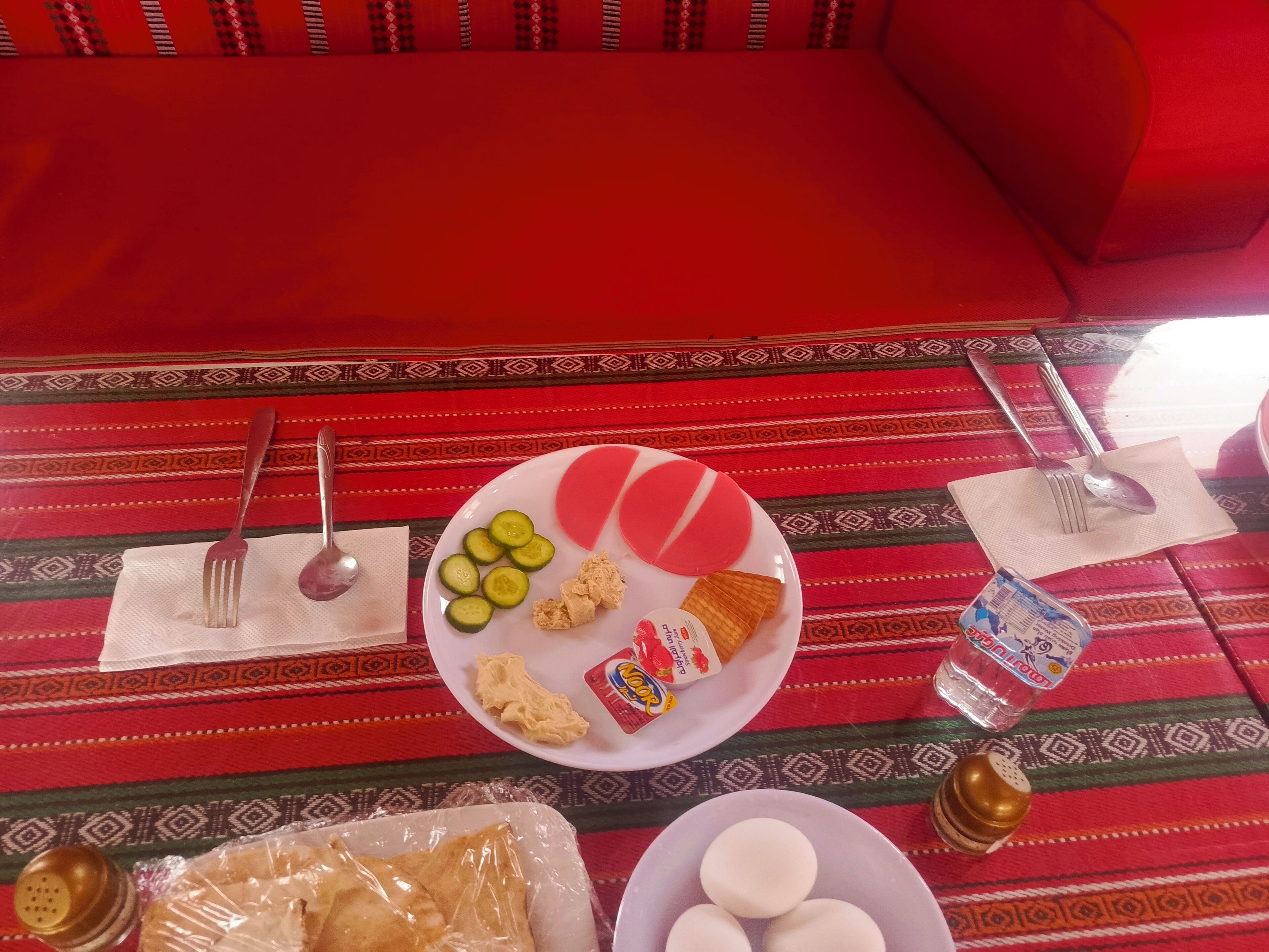A table set with a plate of sliced cucumbers, bread, and a small container of butter, along with a bottle of water and a salt shaker.