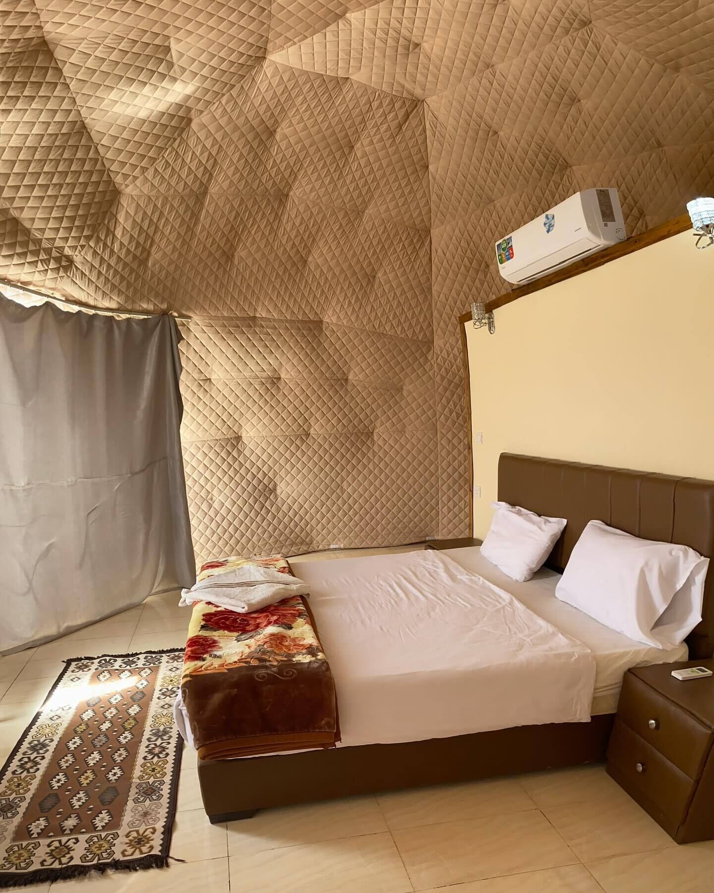 A neatly made bed with a brown headboard, a bedside table with a lamp, and a patterned rug on the floor.