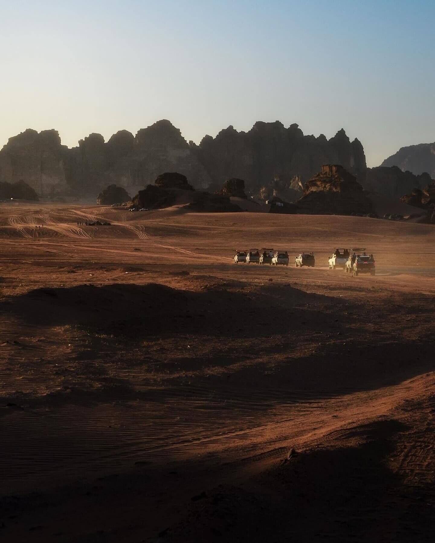 A desert landscape with a group of vehicles and people in the foreground, and rocky formations in the background.