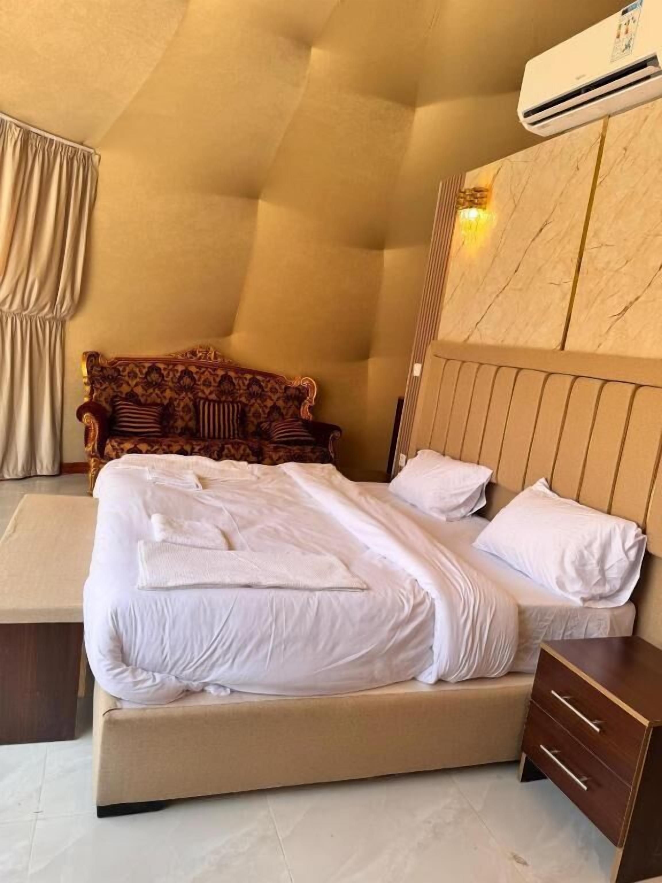 A hotel room with a large bed, a wooden headboard, a nightstand, and an air conditioning unit on the wall.