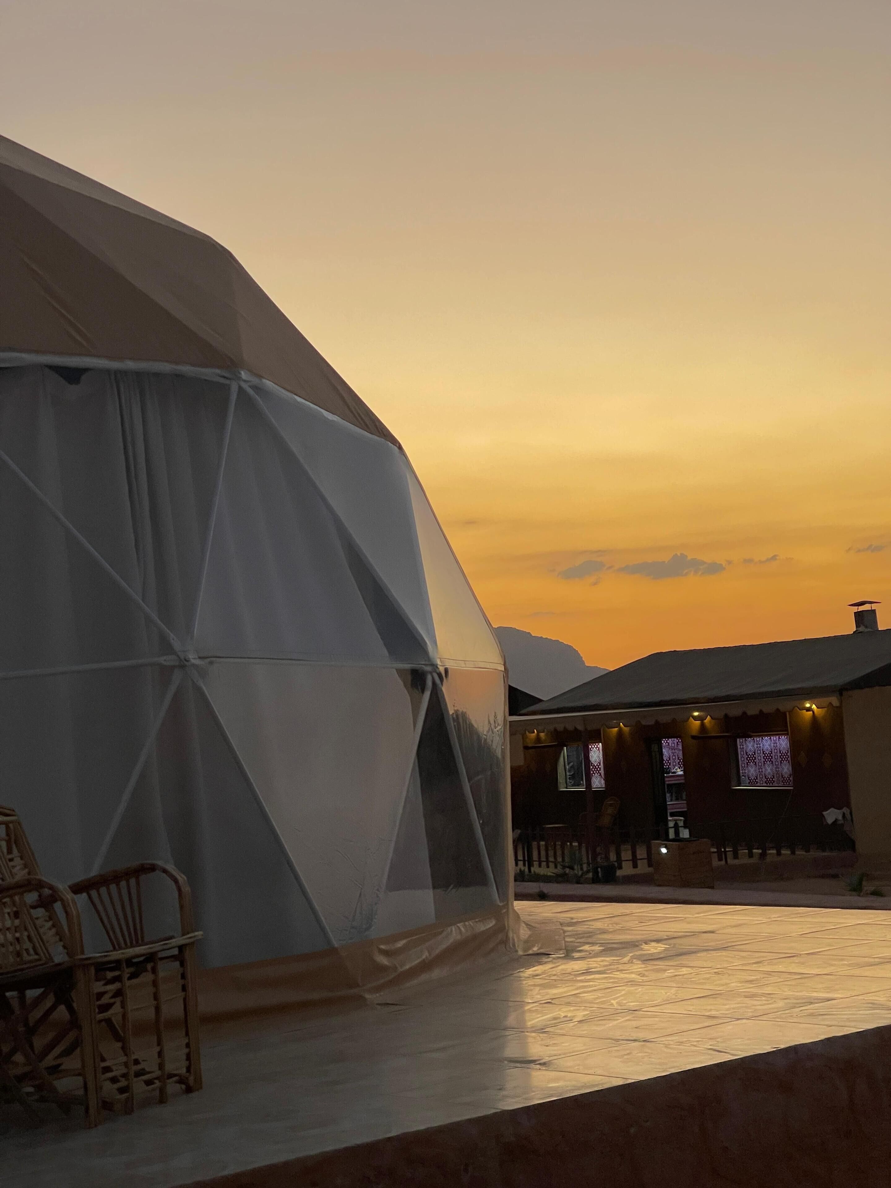 A large geodesic dome structure with a covered patio area, a wooden chair, and a building with windows in the background.