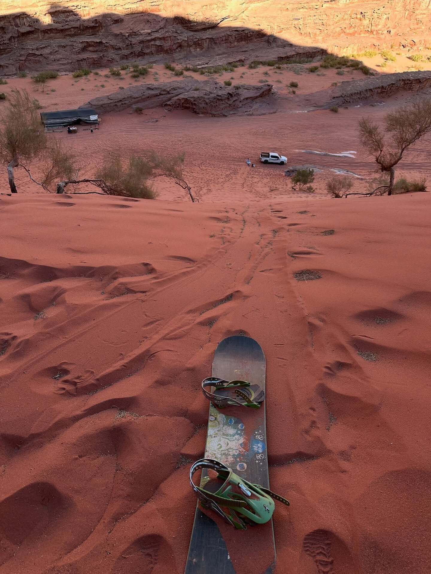 A snowboard on a red sandy surface with footprints, a vehicle, and sparse vegetation in the background.