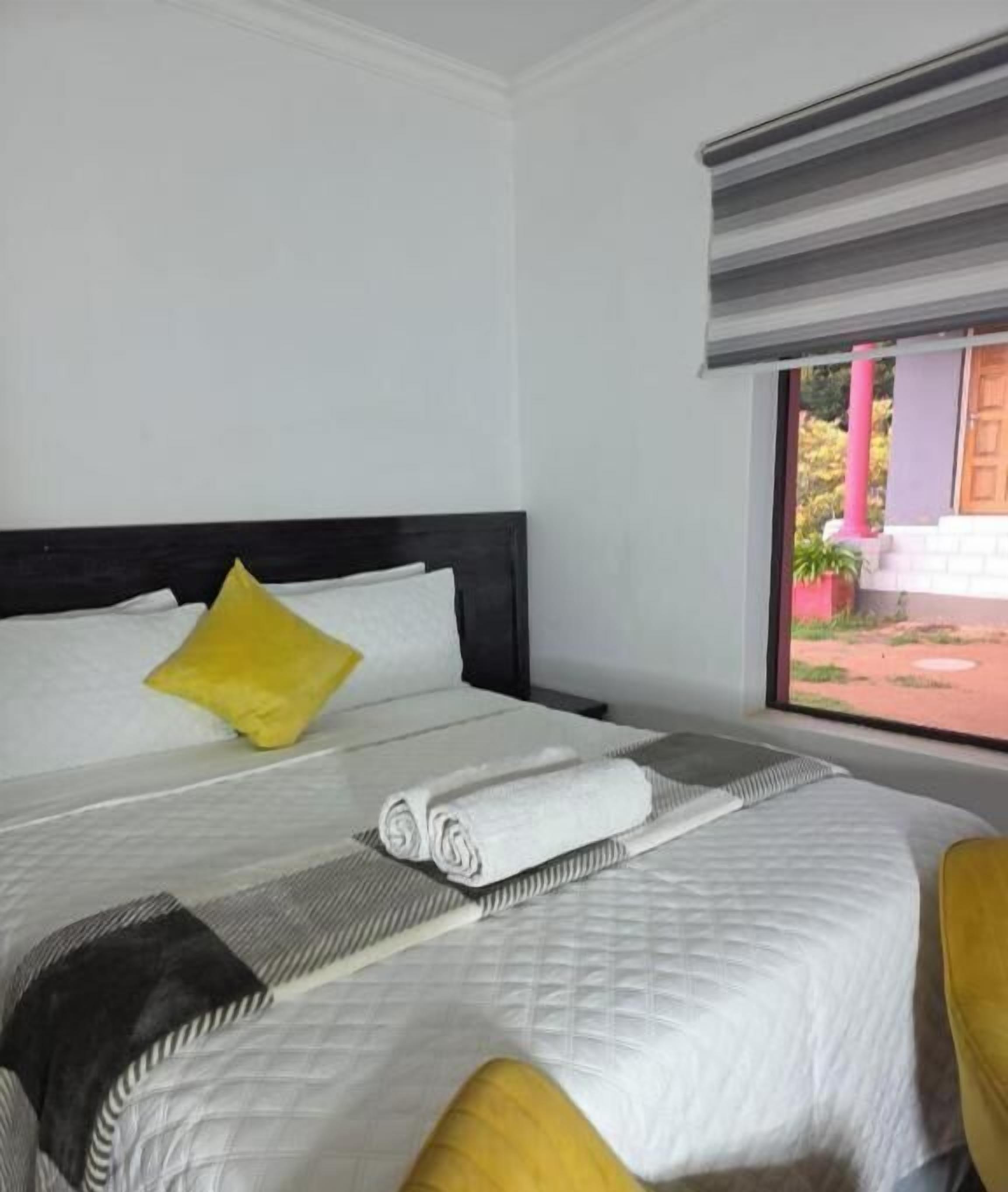 A neatly made bed with white linens and a rolled towel, a yellow pillow, and a striped pillow on a white wall.