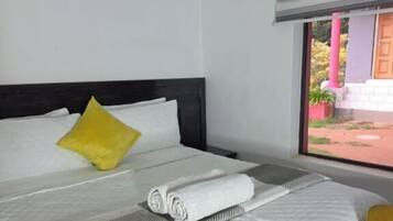 A neatly made bed with white linens and a rolled towel, a yellow pillow, and a striped pillow on a white wall.