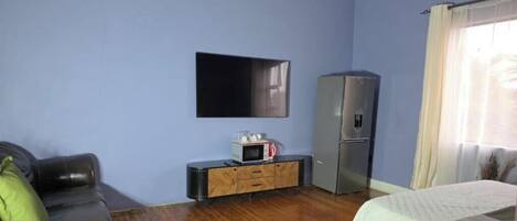 Una habitación con televisión de pantalla plana montada en la pared, refrigerador, microondas y un armario de madera.