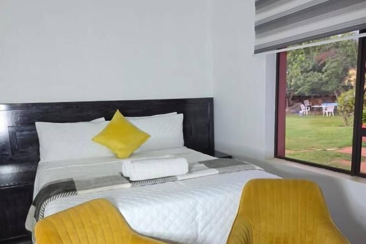 Ein Schlafzimmer mit einem Bett, zwei gelben Stühlen, einem Fenster mit Blick auf einen Garten und einem dunklen Kopfteil.