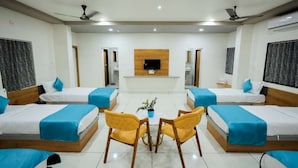 Habitación de hotel con cuatro camas dispuestas en forma de U, una zona de estar central con dos sillones, un televisor de pantalla plana montado en la pared y ventiladores de techo.