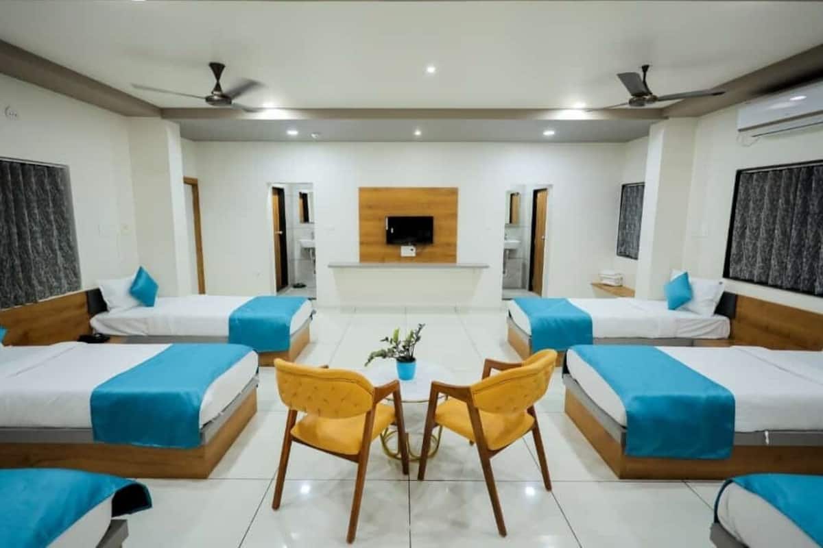 A hotel room with four beds arranged in a U-shape, a central seating area with two chairs, a flat-screen TV mounted on the wall, and ceiling fans.