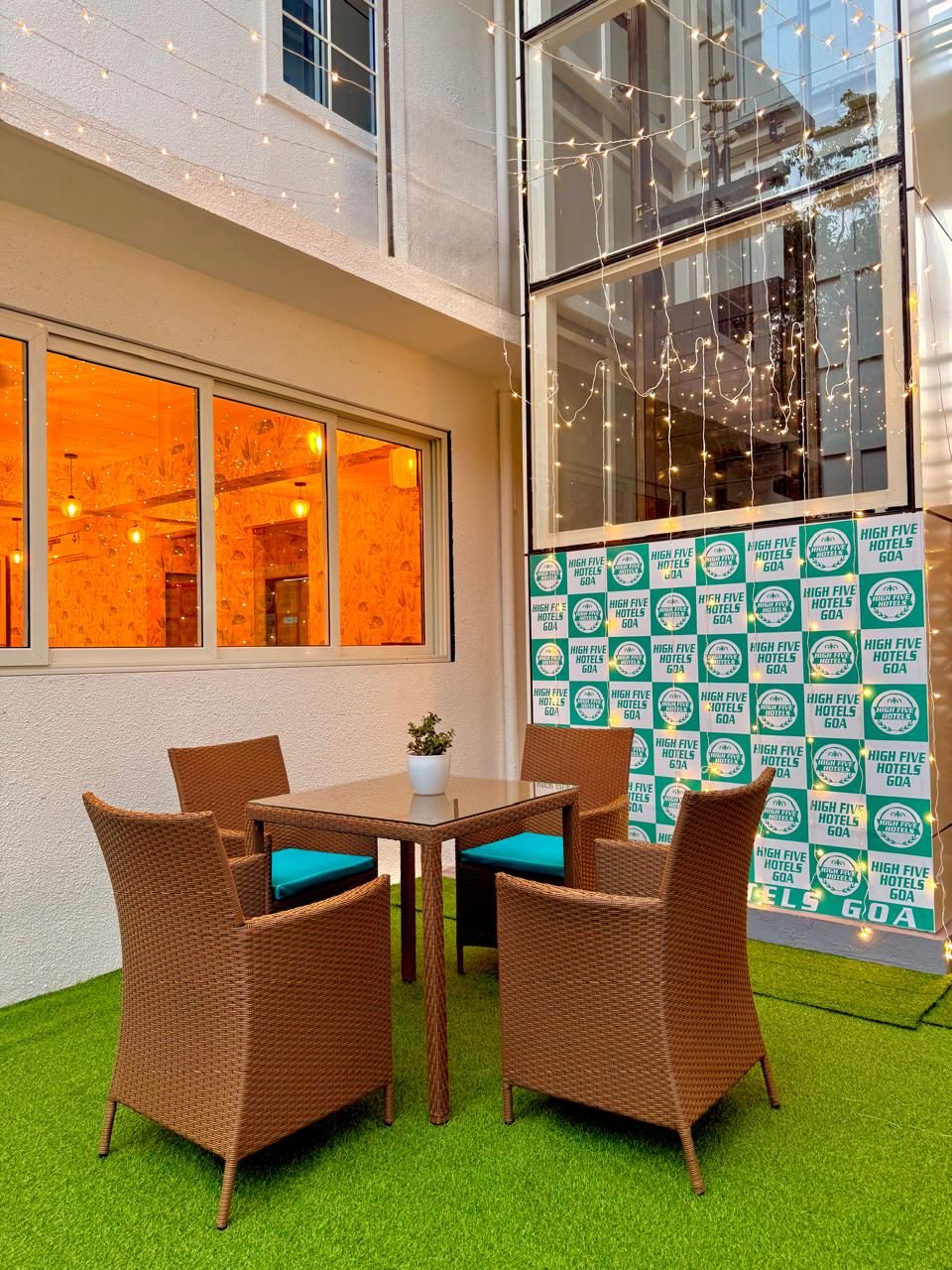 Un espace extérieur aménagé avec des chaises et une table en osier, un mur de verre décoré de guirlandes lumineuses, et un mur orné de plusieurs logos de la chaîne d’hôtels High Five Hotels Goa.