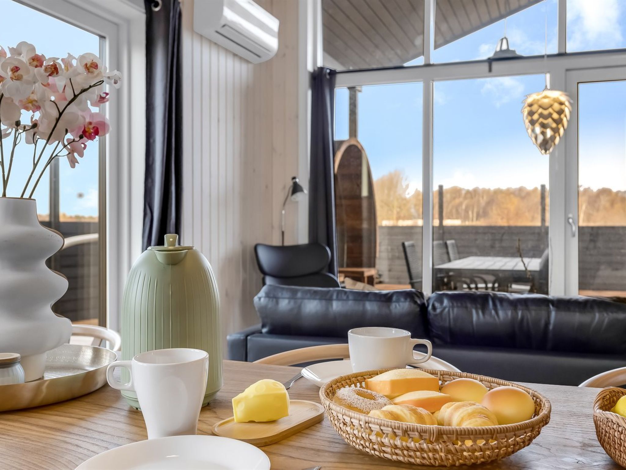 En moderne stue med et stort vindue, en sofa, et spisebord med morgenmadsprodukter og en vase med blomster.