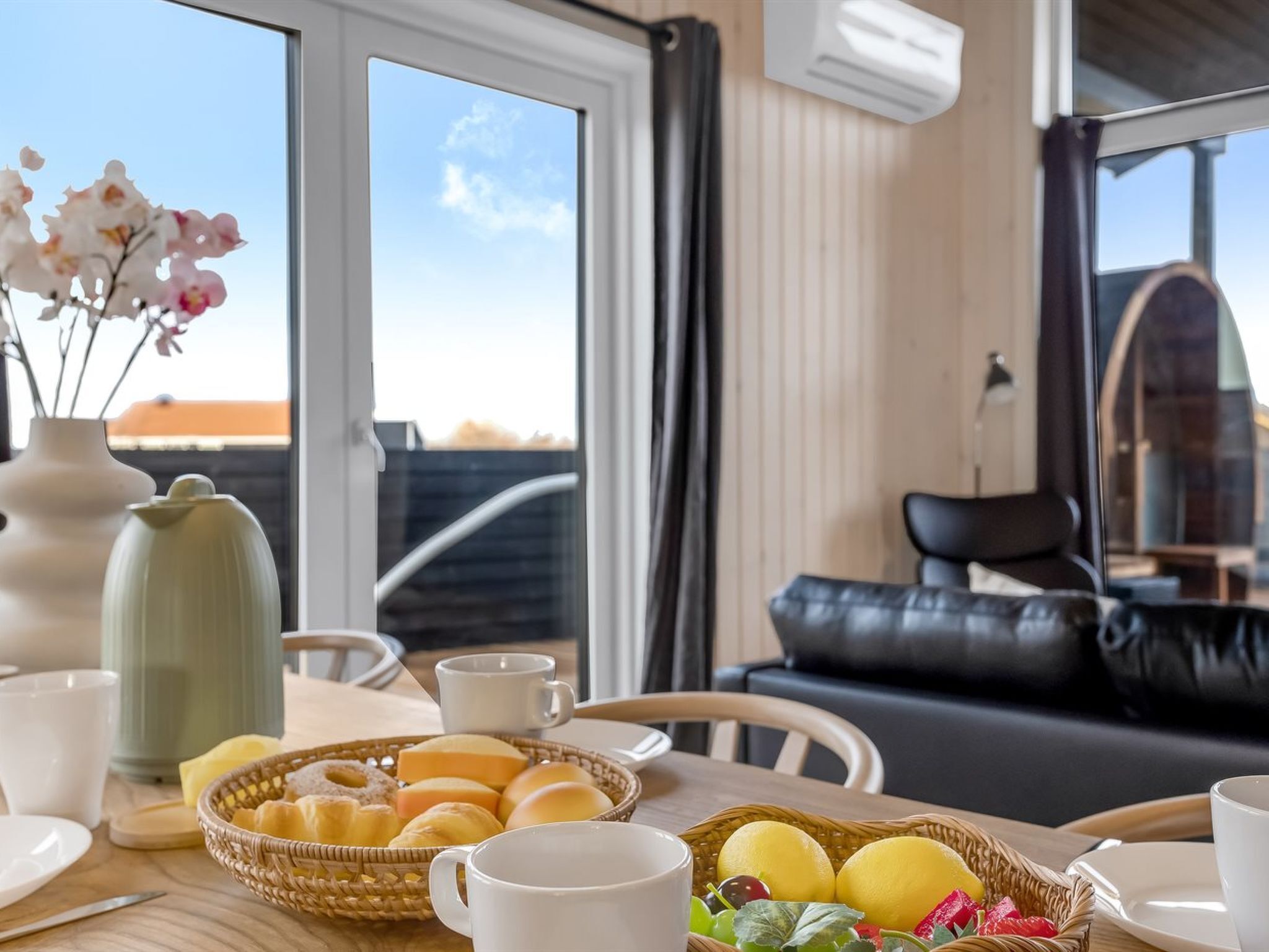A dining area with a table set for breakfast, including pastries and fruits, and a view of the outdoors through large windows.