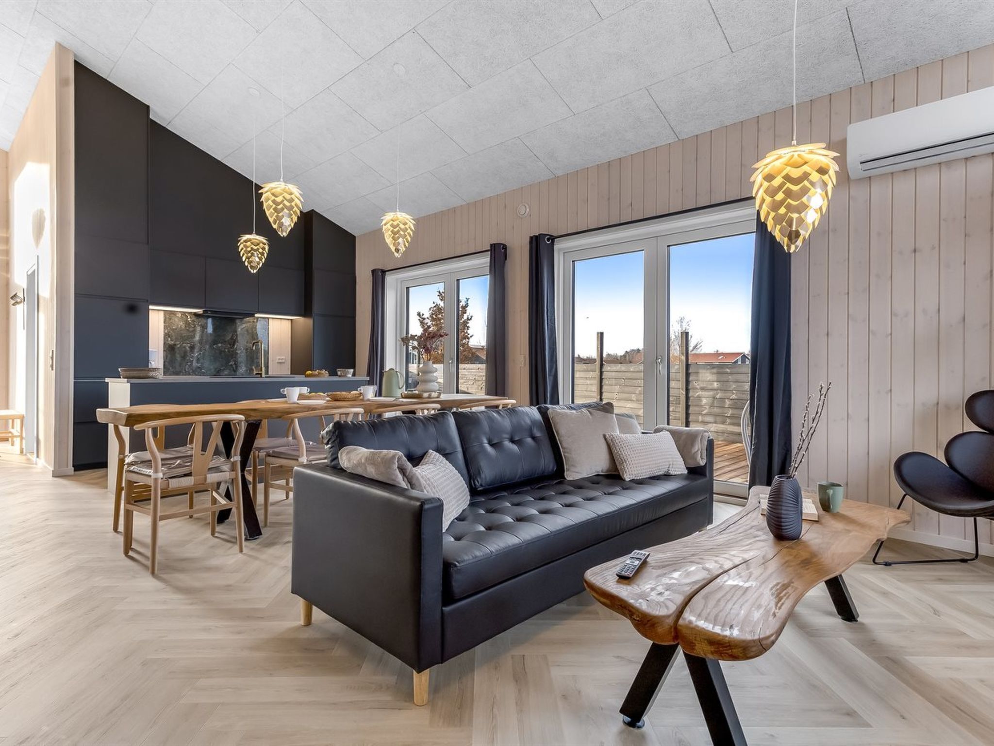 A modern living room with a black leather sofa, wooden dining table, and pendant lights.