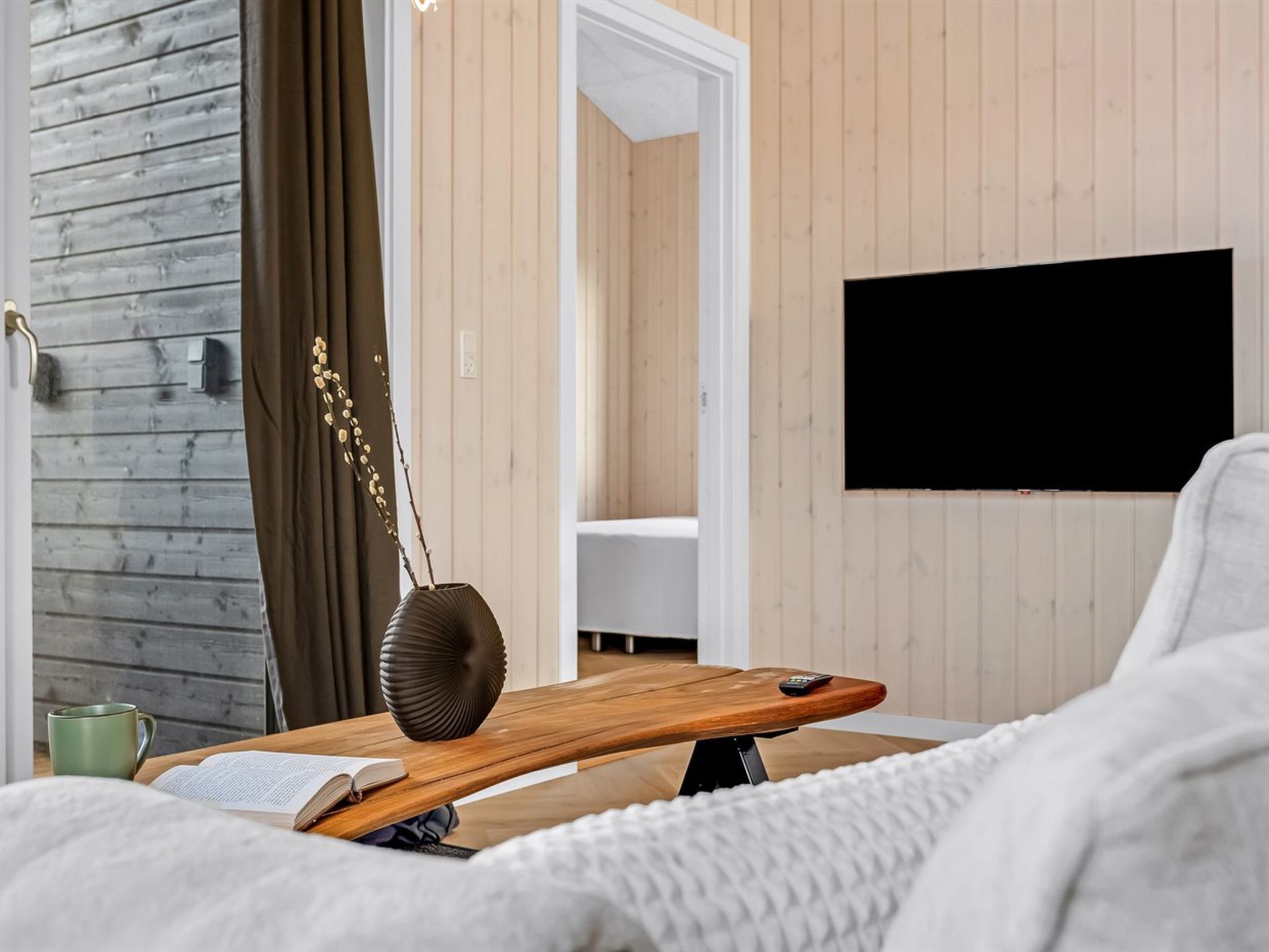 Une chambre moderne avec un lit en bois, une table de chevet sur laquelle se trouve un livre et une tasse verte, un téléviseur fixé au mur et une armoire en bois.