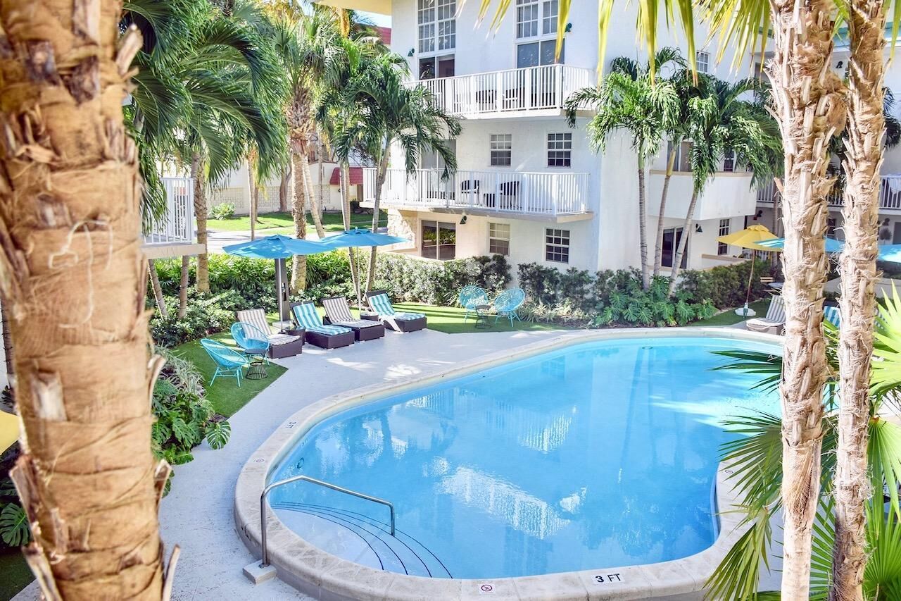 Piscina com espreguiçadeiras e guarda-sóis, cercada por palmeiras e um edifício de vários andares.