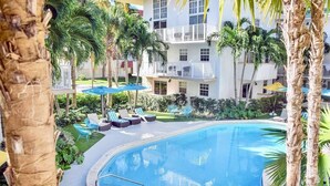 A pool area with lounge chairs and umbrellas, surrounded by palm trees and a multi-story building with balconies. - Relaxed 2BR Coastal Haven with Pool, Minutes from the Beach! - (Key Biscayne)