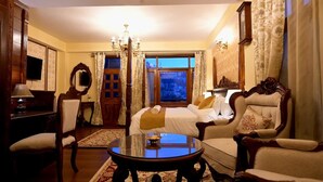 A hotel room with a bed, two armchairs, a round coffee table, a desk with a lamp, a mirror, and a window with curtains. - The Himalayan Heritage (Shimla)