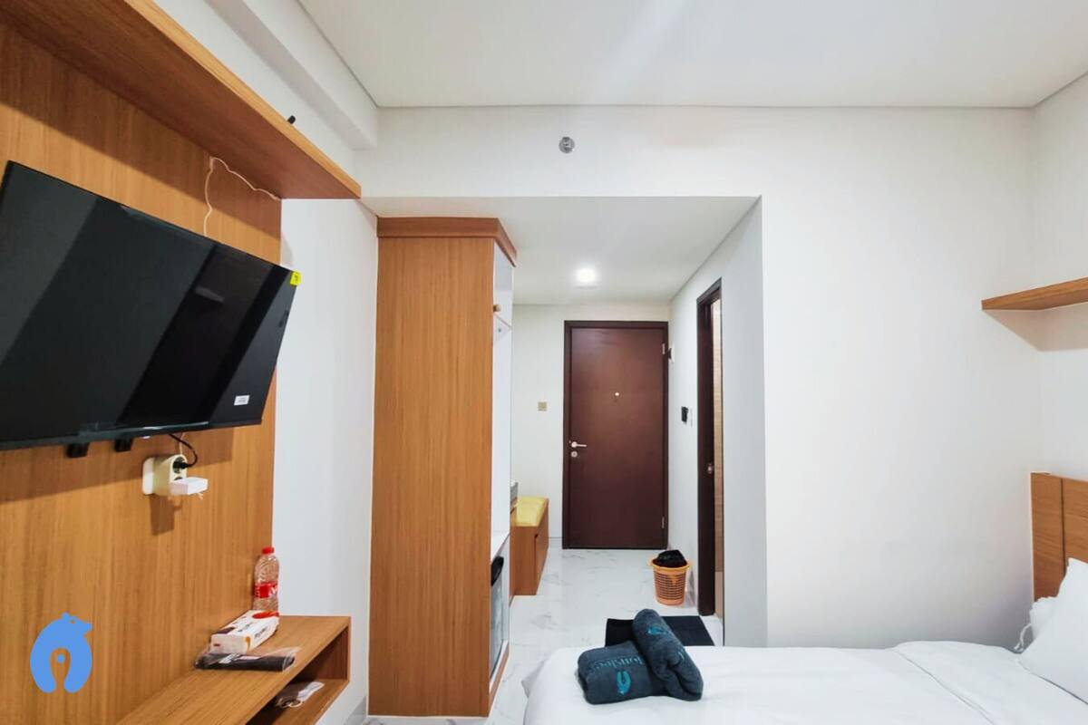 A hotel room with a bed, a TV mounted on the wall, a wooden wardrobe, and a small table with a bottle and a packet on it.