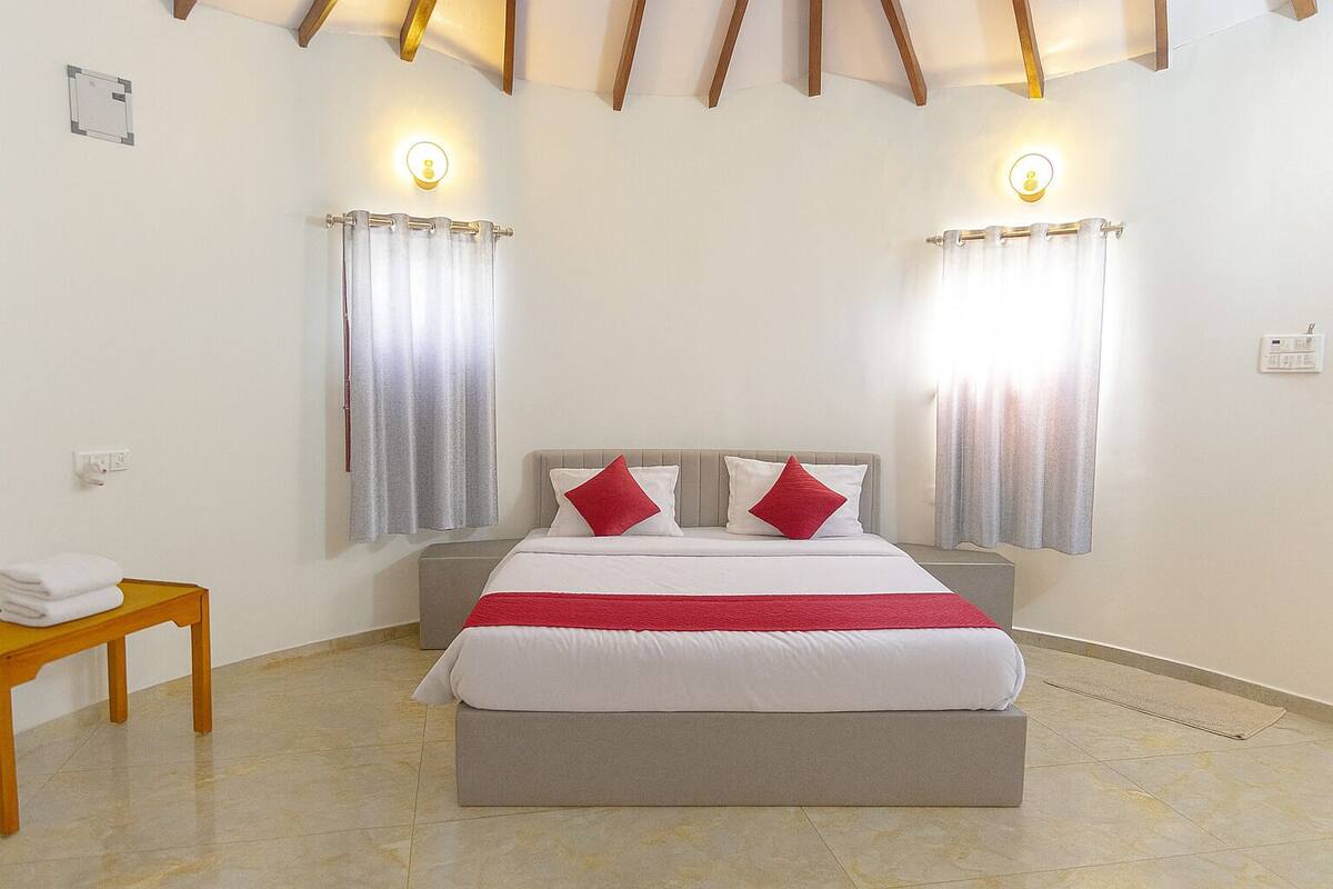 A bedroom with a bed, two bedside tables, a wooden bench, and two windows with curtains.