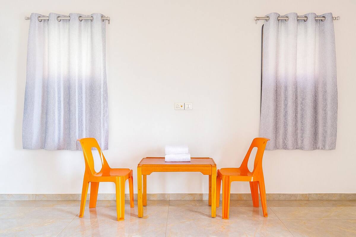 A room with two orange chairs and a small table, a light switch on the wall, and a curtain on the window.