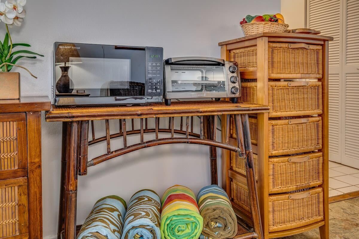 A kitchen area with a microwave and toaster on a wooden shelf, a wicker basket on the counter, and rolled-up towels below.