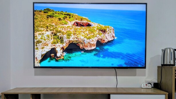 A flat-screen TV mounted on a wall, displaying a coastal landscape. - Private Deluxe Studio - Cozy Chic & Fully Equipped (muscat)