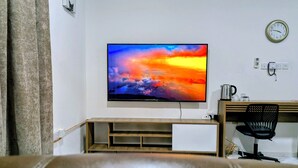 A living room with a flat-screen TV mounted on the wall, a wooden desk, a black office chair, and a clock on the wall. - Private Deluxe Studio - Cozy Chic & Fully Equipped (muscat)