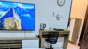 Un salon moderne avec un canapé en cuir, une table basse blanche, un téléviseur affichant un paysage montagneux, un bureau en bois avec une chaise et une horloge au mur.