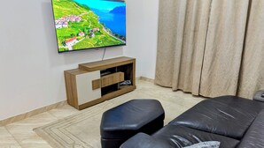 Ett vardagsrum med en platt-TV som är monterad på väggen, ett TV-bord i trä med några lådor, en svart lädersoffa och en mönstrad gardin.