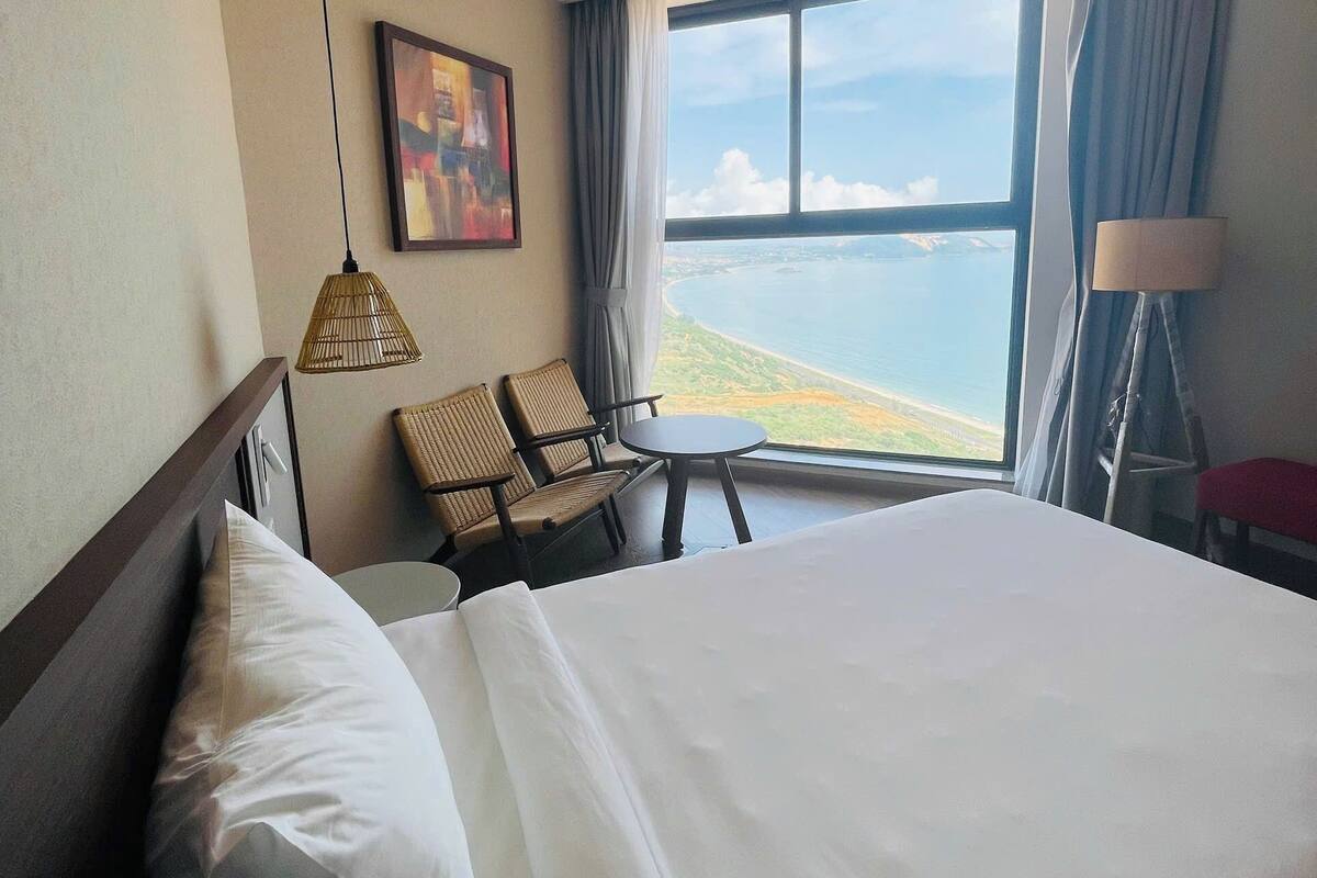 Een hotelkamer met een groot bed, een eettafel, stoelen, een hanglamp, een schilderij en uitzicht op zee.