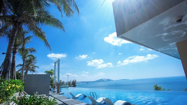 A poolside area with lounge chairs, a palm tree, and a view of the sea.