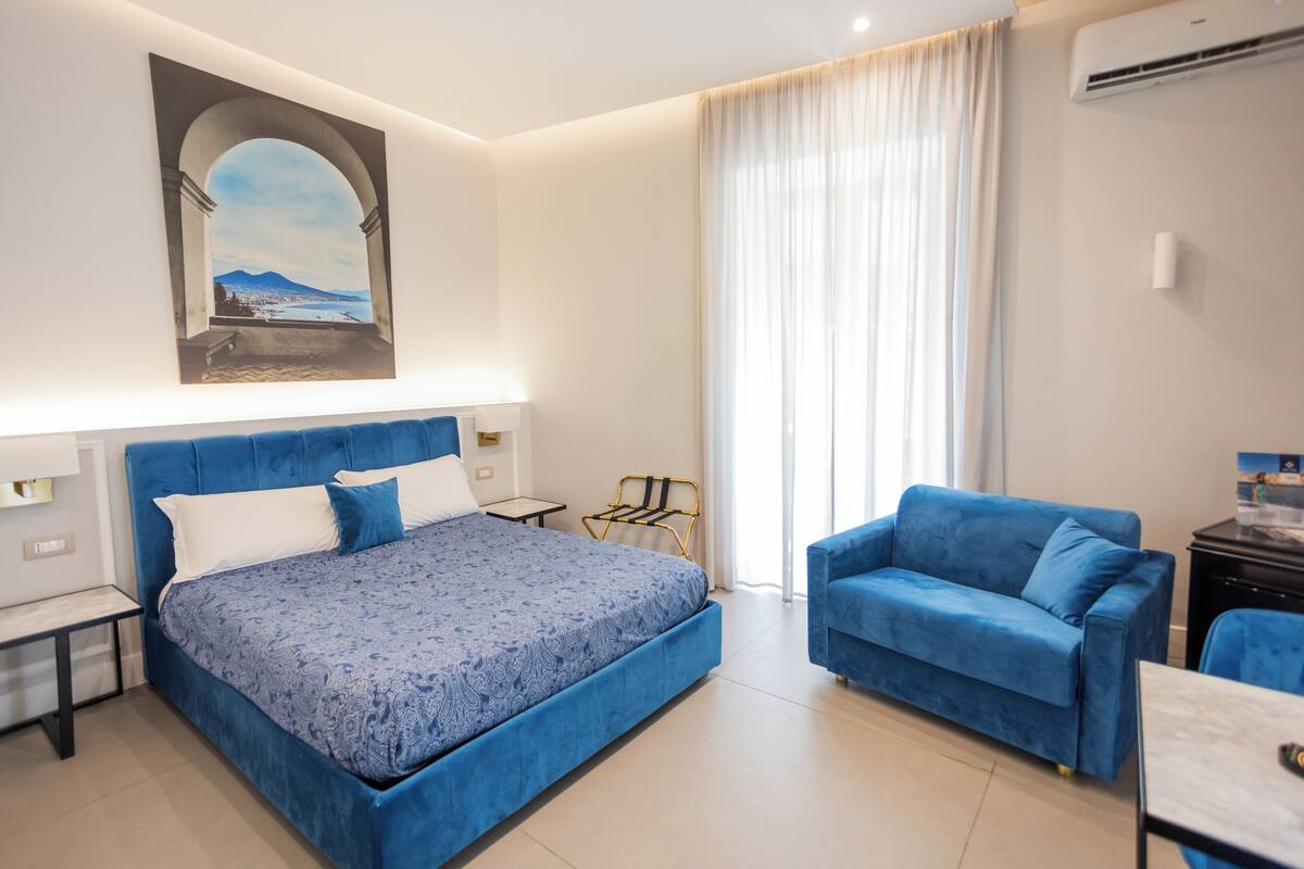 Habitación de hotel moderna con una cama grande, un sofá azul, una mesita y un cuadro de un paisaje.