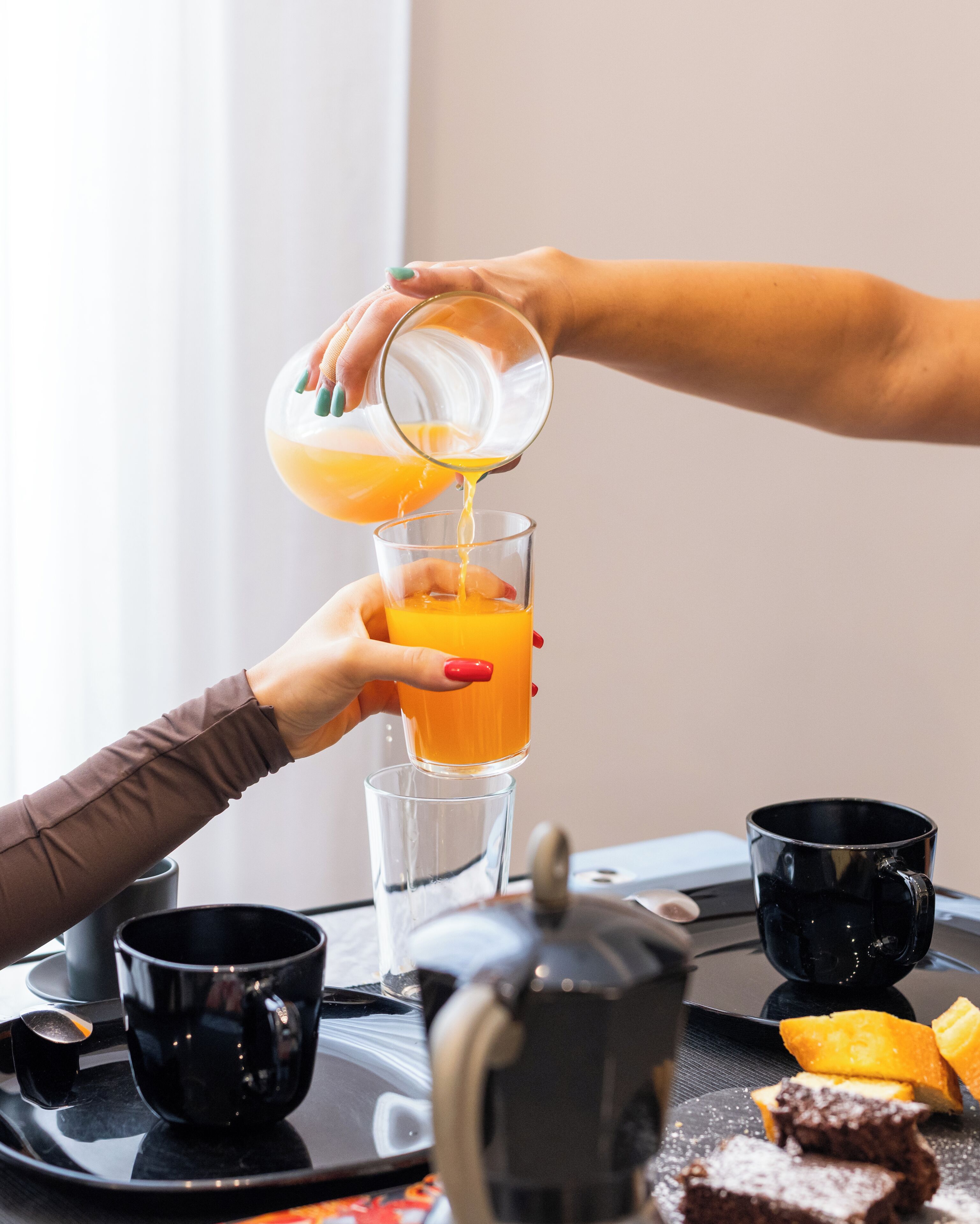 A breakfast tray with a glass of orange juice, two black mugs, a teapot, and a slice of cake.