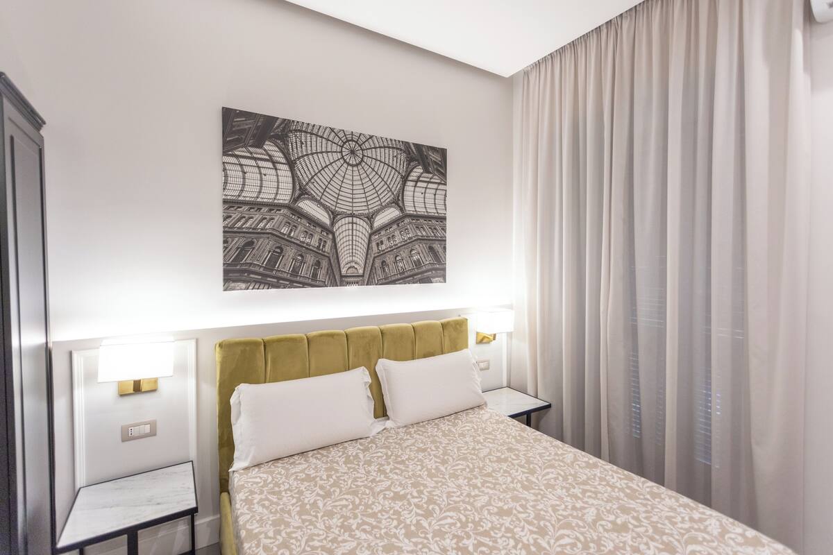 Habitación de hotel con una cama grande, cabecera, dos mesitas de noche, una lámpara de pared y un amplio ventanal con cortinas.