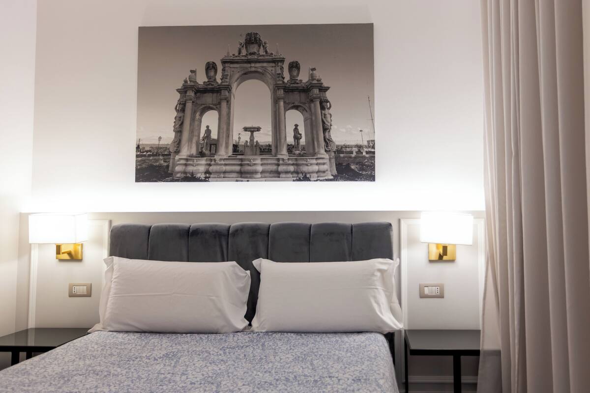 Un dormitorio con cama, dos lámparas de noche, un cuadro colgado en la pared de un arco y una ventana con cortinas.