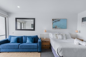 A modern bedroom with a blue sofa, a bed with white bedding, a nightstand with a lamp, and a large mirror on the wall. - Bay View - Ocean Nest (Lorne)