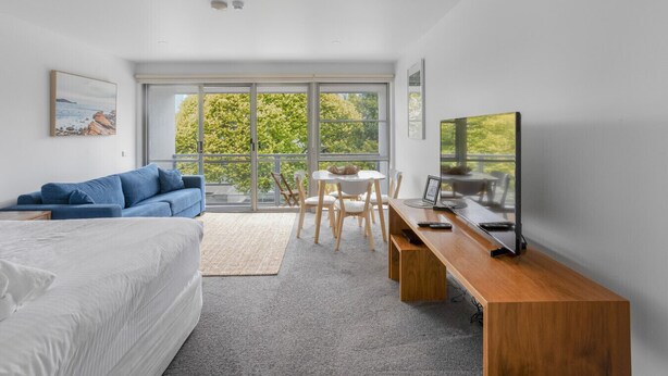 A modern bedroom with a large bed, a blue sofa, a dining table with chairs, a TV, and large windows offering a view of trees and a building.