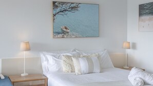 A neatly made bed with white linens and a rolled towel, two bedside lamps, a wooden nightstand, and a painting of a tree by a body of water.