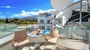 A balcony with two wicker chairs, a small round table with a fruit bowl, and a view of a modern building with a staircase. - Pnoe Beachside Studio (Georgioupoli)