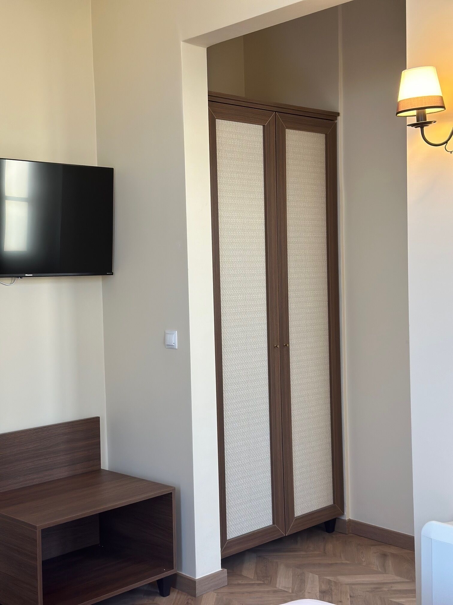 A room with a flat-screen TV mounted on the wall, a wooden bench, a wardrobe with a mirror, and a wall-mounted lamp.
