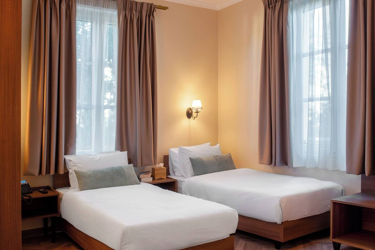 A hotel room with two beds, a bedside table with a lamp, and a window with curtains.