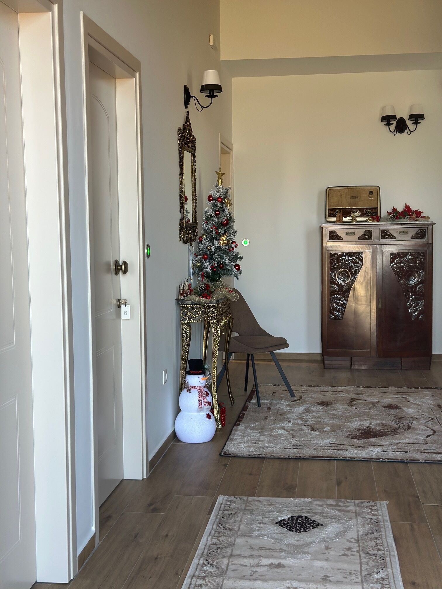 A hallway with a white door, a small Christmas tree, a snowman decoration, a vintage radio, and a cabinet with decorative items.