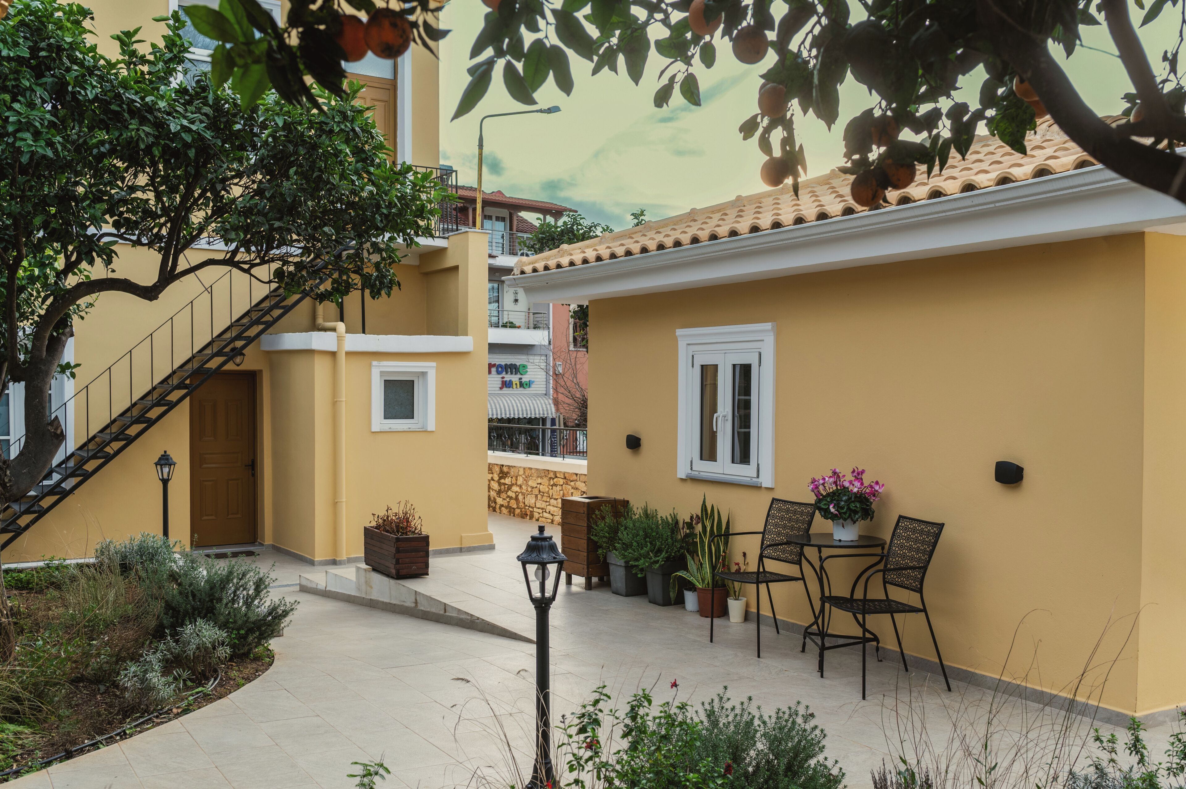 A yellow building with a staircase, a small patio with chairs and potted plants, and a stone wall in the background.