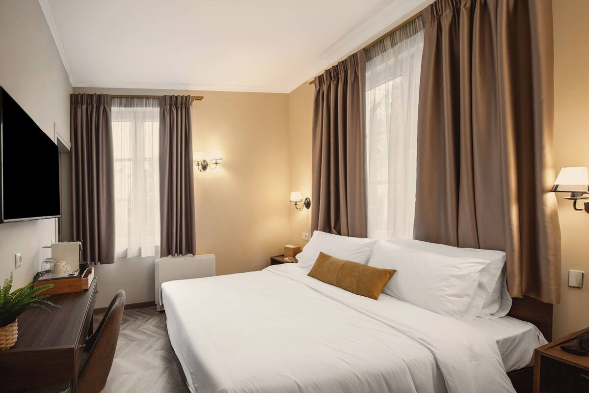 A hotel room with a bed, a desk, a chair, a flat-screen TV, and a window with curtains.