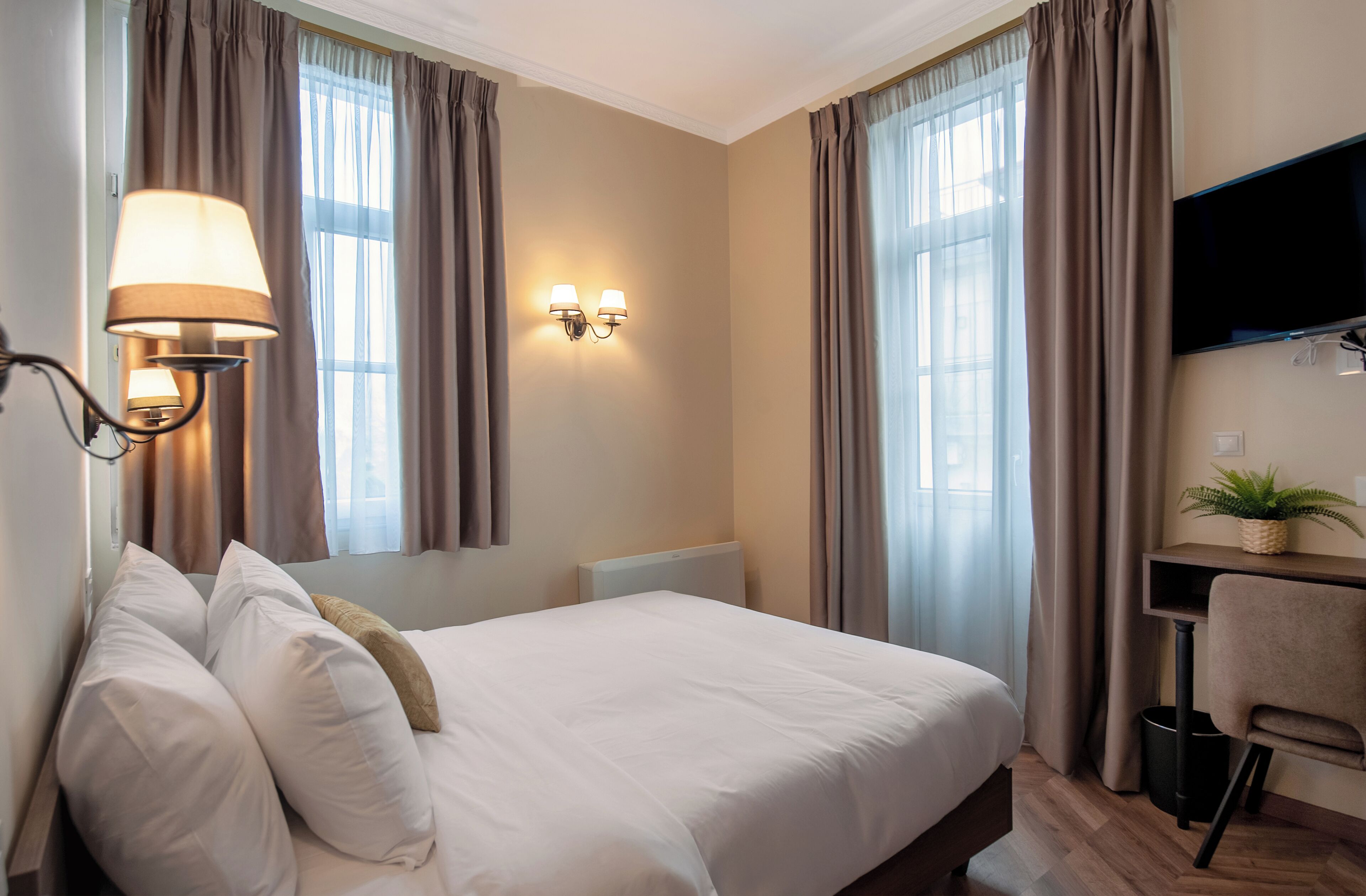 A hotel room with a bed, a desk, a chair, a TV, and a wall-mounted lamp.