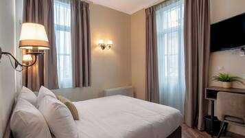A hotel room with a bed, a desk, a chair, a TV, and a wall-mounted lamp.