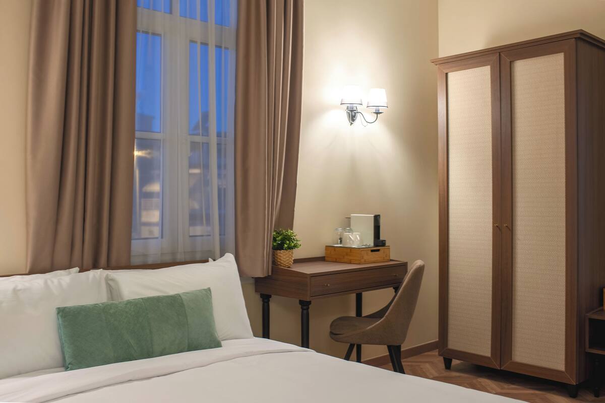 A hotel room with a bed, a desk with a lamp, a wardrobe, and a window with curtains.