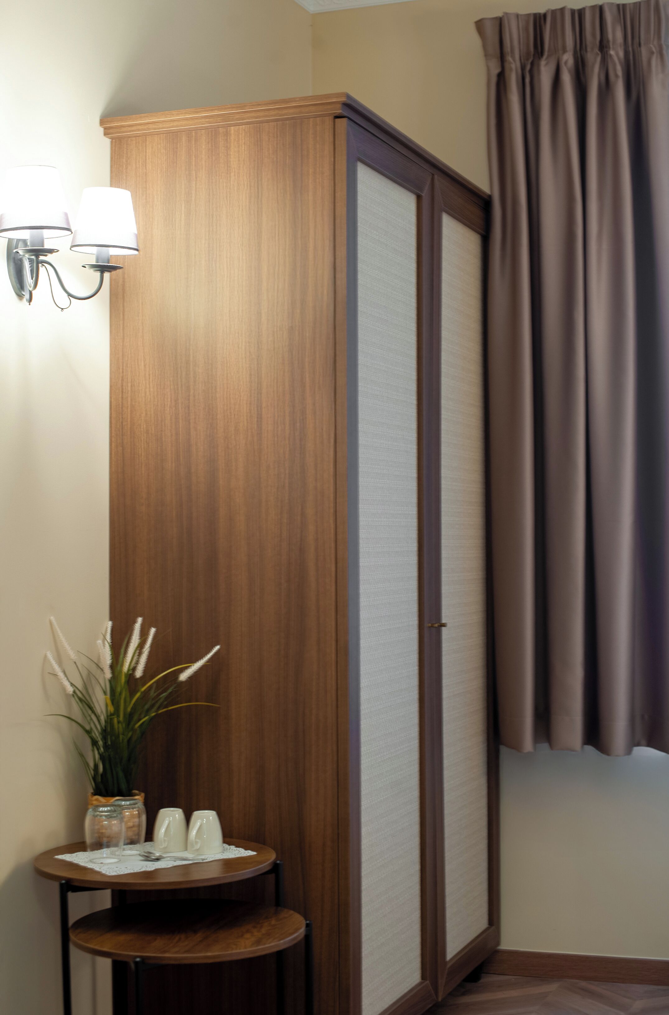 A room with a wooden wardrobe, a small table with a plant, salt and pepper shakers, and a wall-mounted light fixture.