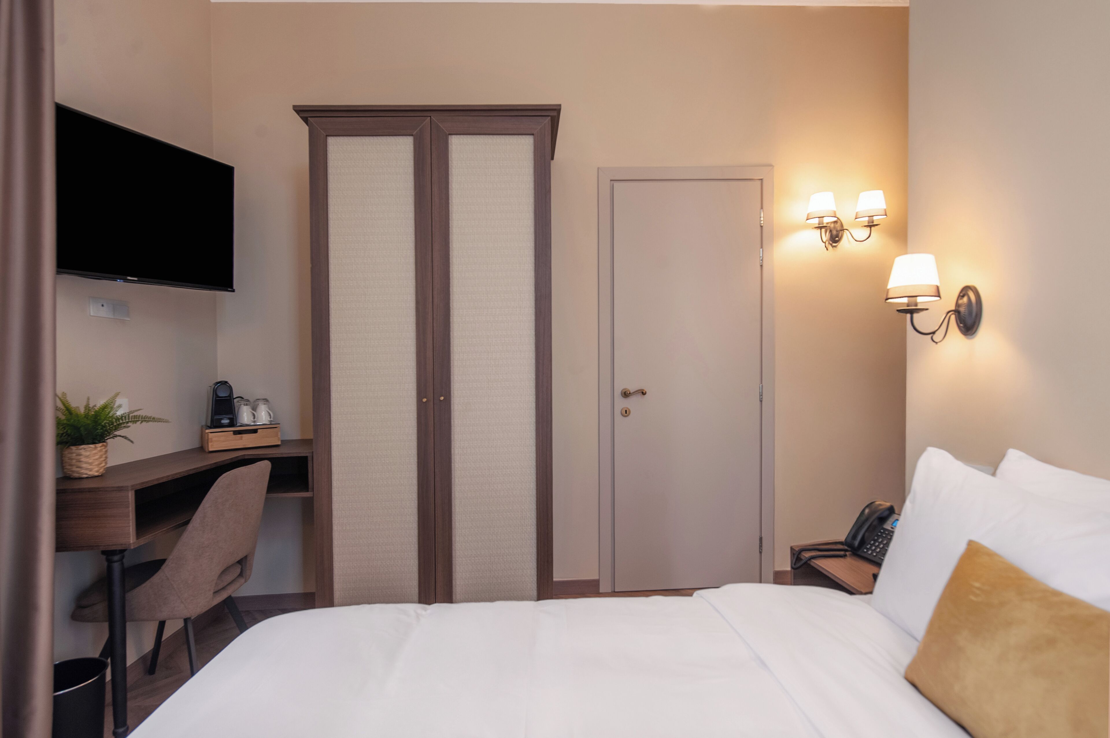 A hotel room with a bed, a desk with a chair, a TV mounted on the wall, a wardrobe, and a nightstand with a lamp.