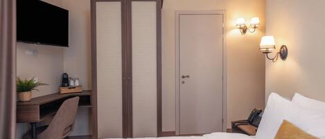 A hotel room with a bed, a desk with a chair, a TV mounted on the wall, a wardrobe, and a nightstand with a lamp.